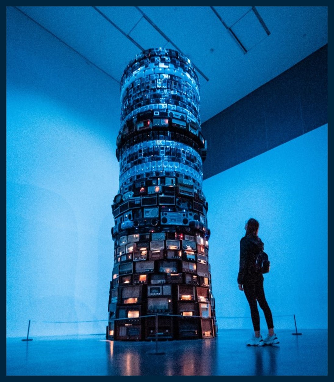A column built of different electronic devices such as radios and televisions. A woman is looking up at the column.
