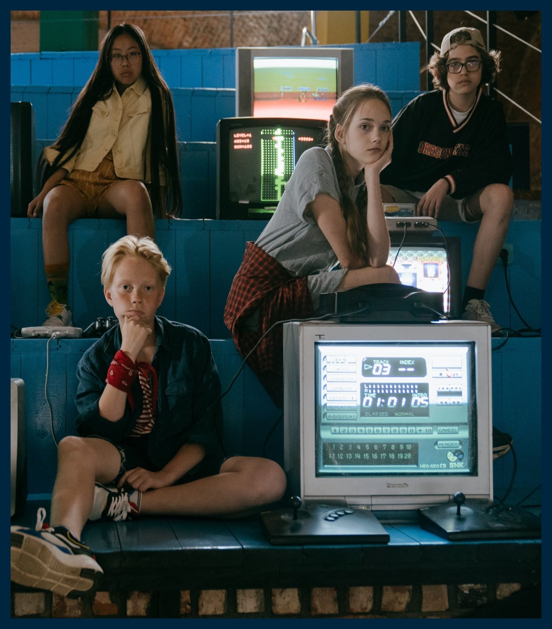 A group of young people looking at the camera. There are old televisions between them with different programs on.