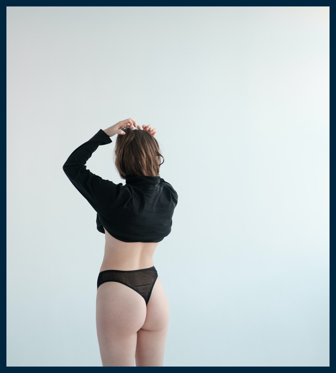 A woman standing in a white room, wearing a black shirt and underpants.